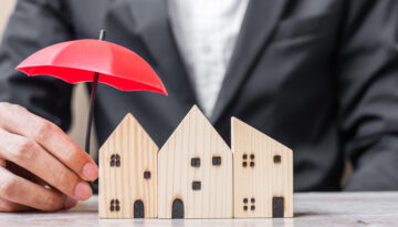 Businessman hand holding red Umbrella cover wooden Home model on table office. Property insurance and real estate concepts