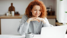 Female freelancer having video conference with her employer discussing new project details, listening to him attentively in headphones smiling at web camera, putting folded hands under chin