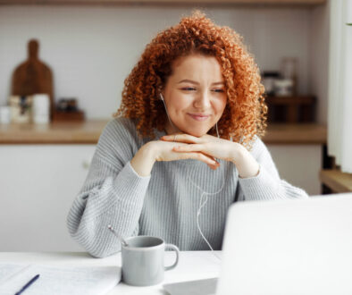 Female freelancer having video conference with her employer discussing new project details, listening to him attentively in headphones smiling at web camera, putting folded hands under chin