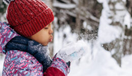 Happy little African-American girl in a red hat and overalls blowing snow off the hand.Winter, Christmas and Happy New Year concept.