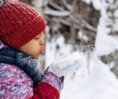 Happy little African-American girl in a red hat and overalls blowing snow off the hand.Winter, Christmas and Happy New Year concept.